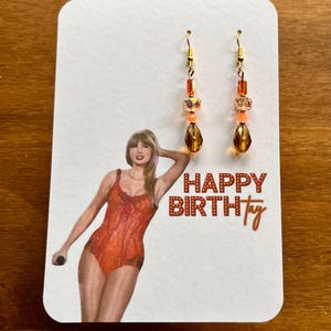 May include: A pair of dangle earrings with gold-colored hooks. The earrings feature a combination of orange and amber-colored beads and teardrop-shaped pendants. The earrings are displayed on a white card with a graphic and the text "HAPPY BIRTHtay".