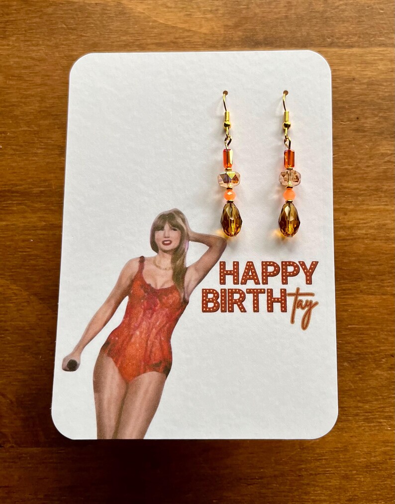 May include: A pair of dangle earrings with gold-colored hooks. The earrings feature a combination of orange and amber-colored beads and teardrop-shaped pendants. The earrings are displayed on a white card with a graphic and the text "HAPPY BIRTHtay".