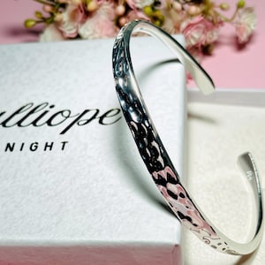May include: A silver-toned metal cuff bracelet featuring a hammered, reflective surface texture. The bracelet is positioned next to a white rectangular box with the black printed text Calliope MIDNIGHT visible on the top surface of the box.