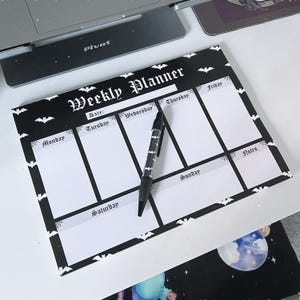May include: A black and white weekly planner pad featuring a bat and spiderweb pattern with the text Weekly Planner. The planner has sections for each day of the week and a notes section. A black pen with a white bat pattern rests on the planner.