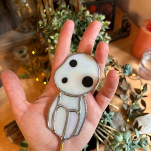 May include: A white stained glass figurine of a character from the film "My Neighbor Totoro" with black eyes. The figurine is attached to a gold metal rod.