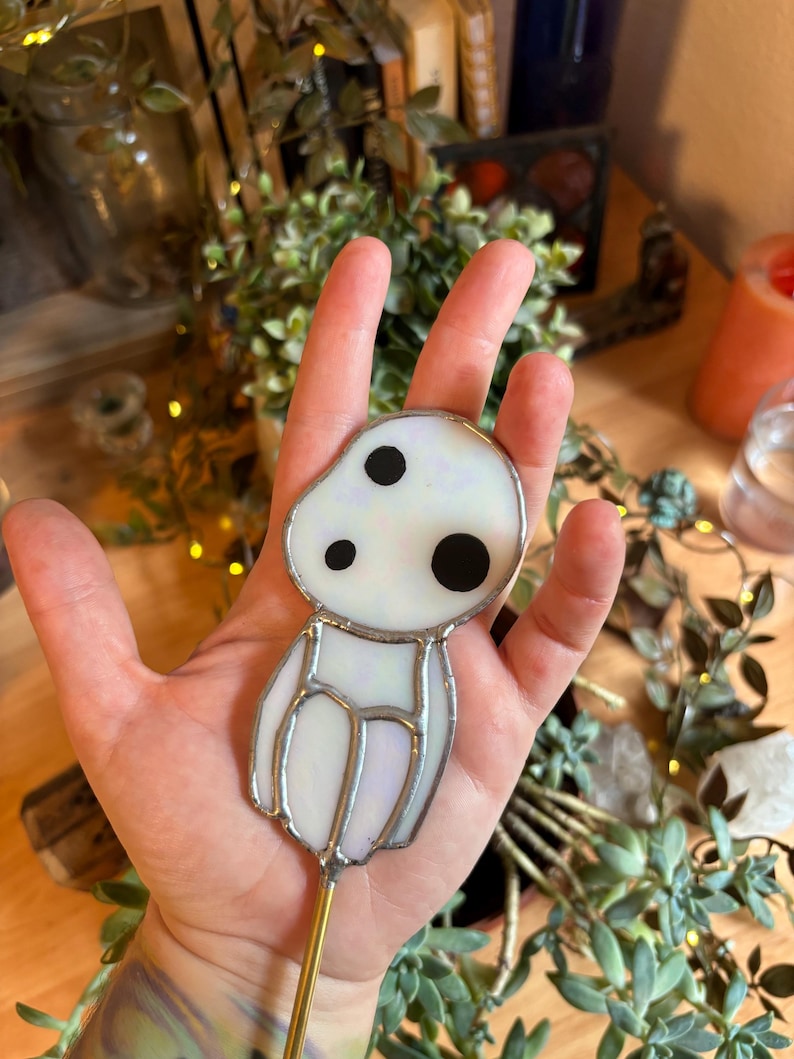 May include: A white stained glass figurine of a character from the film "My Neighbor Totoro" with black eyes. The figurine is attached to a gold metal rod.