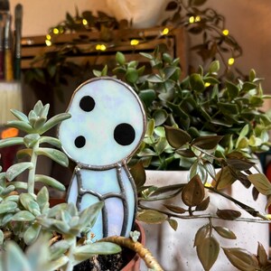 May include: A stained glass figurine of a white, translucent character with black eyes. The figurine is sitting in a terracotta pot with a succulent plant.