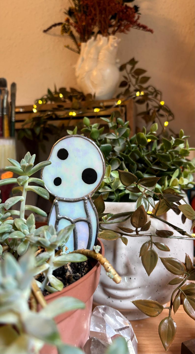 May include: A stained glass figurine of a white, translucent character with black eyes. The figurine is sitting in a terracotta pot with a succulent plant.