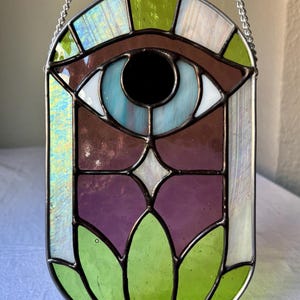 Puede incluir: Un ojo de vidrio coloreado con un iris azul y una pupila negra, rodeado de un fondo marrón y morado. El ojo está colocado en un marco verde y blanco con una cadena para colgar.
