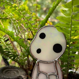 May include: A white stained glass figurine of a character with three black dots for eyes. The figurine is sitting in a pot with green ferns and a grey fossil.