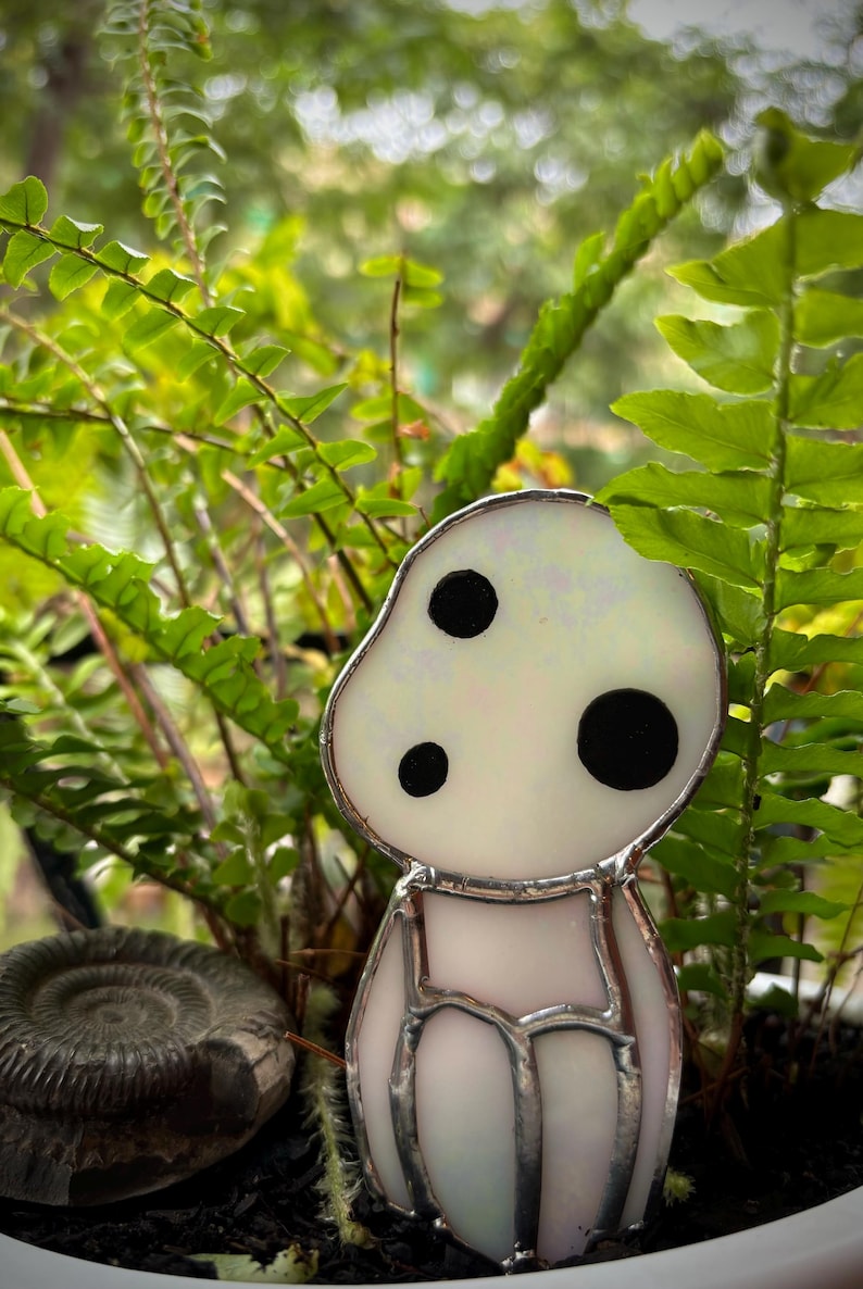 May include: A white stained glass figurine of a character with three black dots for eyes. The figurine is sitting in a pot with green ferns and a grey fossil.