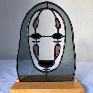 Puede incluir: Una representación en vitral de Sin Cara de la película animada El viaje de Chihiro. El vitral está colocado sobre un soporte de madera. La cara es blanca con ojos negros y una boca negra. El cabello es de color azul oscuro.
