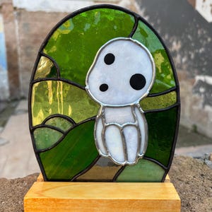May include: A stained glass depiction of a white, round, cartoon character with black eyes, set against a green and gold background. The stained glass piece is mounted on a wooden stand.