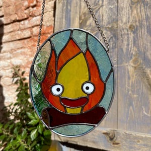 May include: A stained glass window hanging from a chain, depicting a cartoon fire with a smiling face. The fire is orange, yellow, and red, with white eyes and a red mouth.