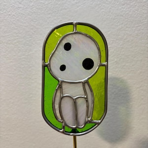 May include: A stained glass suncatcher featuring a white, cartoon-like character with three black eyes. The character is sitting on a green background.