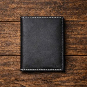 May include: A rectangular black leather wallet featuring a smooth, matte finish and contrasting white stitching along the edges. The wallet is oriented vertically, showcasing a clean, simple design with a visible seam running along the perimeter.