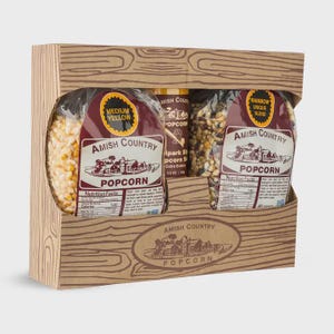 May include: A wooden gift box containing Amish Country popcorn. The box has a wood grain design and holds two bags of popcorn and a small container. One bag is labeled "Medium Yellow" and the other "Rainbow Unique Blend."