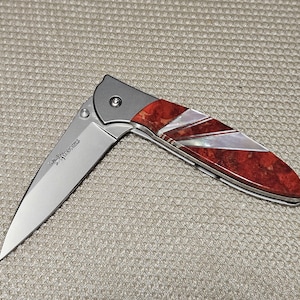 May include: A folding knife with a silver blade and a handle featuring red and white inlaid design. The handle has a silver bolster and a decorative pattern. The blade is partially open, revealing the knife's sharp edge.