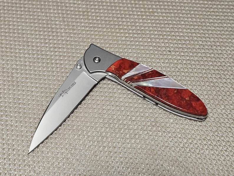 May include: A folding knife with a silver blade and a handle featuring red and white inlaid design. The handle has a silver bolster and a decorative pattern. The blade is partially open, revealing the knife's sharp edge.