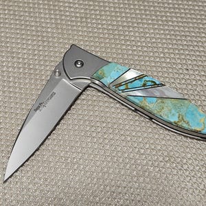 Puede incluir: Una navaja plegable parcialmente abierta con una hoja plateada y un mango con incrustaciones de turquesa y n&aacute;car. La navaja tiene una virola plateada y una bisagra visible. La hoja tiene el texto "Serge Panchenko Knives".