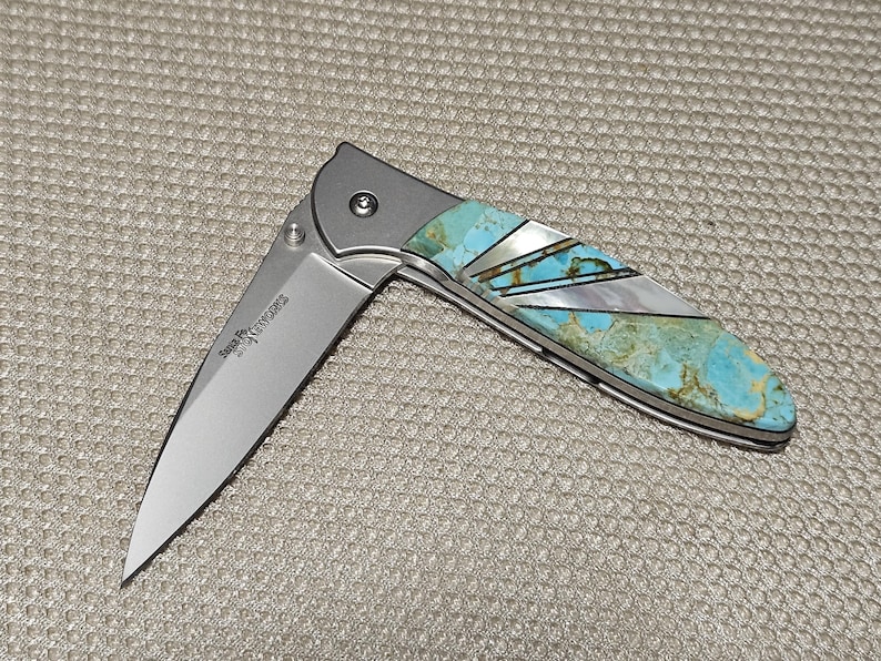 Puede incluir: Una navaja plegable parcialmente abierta con una hoja plateada y un mango con incrustaciones de turquesa y n&aacute;car. La navaja tiene una virola plateada y una bisagra visible. La hoja tiene el texto "Serge Panchenko Knives".