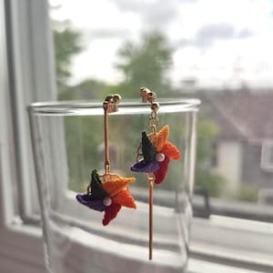 May include: A pair of handmade, colorful pinwheel earrings. Each earring features a pinwheel design with red, orange, yellow, green, and purple crochet petals, accented with a small white bead in the center. The earrings have gold-colored posts and long, slender gold-colored bars.