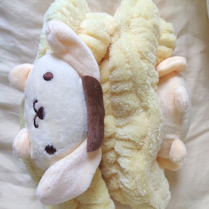 May include: A pair of yellow plush scrunchies with a cartoon bunny face. The bunny has a white face, brown ear, and black embroidered details. The scrunchies are made of a soft, textured material.