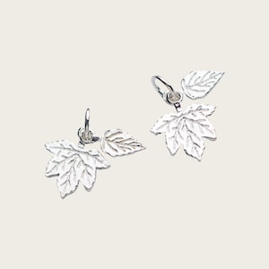 May include: Two silver leaf-shaped charms with detailed veining. Each charm features a larger maple leaf and a smaller leaf, suspended from a small loop. The charms are displayed against a light background, highlighting their metallic sheen.