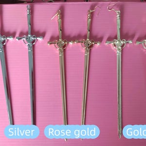 Puede incluir: Pendientes con forma de espada en plata, oro rosa y oro. Los pendientes tienen una empuñadura decorativa y una hoja larga y delgada. Las palabras "Silver", "Rose gold" y "Gold" están escritas en la imagen.