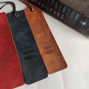 Könnte beinhalten: Drei Leder-Lesezeichen in Rot, Marineblau und Hellbraun. Jedes Lesezeichen hat eine Lederschlaufe und zeigt ein Sternbild-Design und den Text "FROM THE LIBRARY OF" gefolgt von einem Namen. Im Hintergrund ist ein Buch mit dem Titel "SEVENEVES" zu sehen.