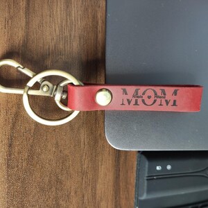 Puede incluir: Llavero de cuero rojo con cierre dorado. El llavero tiene la palabra "MOM" y los nombres "Ariela, Milana" grabados. Un accesorio elegante para llaves o bolsos.