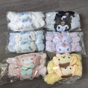 May include: Six plush headbands in clear plastic packaging. The headbands feature cartoon character designs in white, black, light blue, lavender, pink, and yellow. Each headband has a different character design with bow accents.