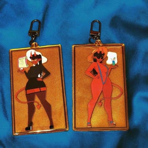 May include: Two rectangular keychains with gold frames and black metal clasps. Each keychain features a cartoon devil woman illustration. One is in a black outfit, the other in a red bikini. Both hold items and have red horns and tails.