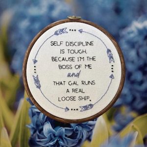 May include: An embroidered hoop featuring the text "SELF DISCIPLINE IS TOUGH, BECAUSE I'M THE BOSS OF ME and THAT GAL RUNS A REAL LOOSE SHIP." The text is in black thread on white fabric, within a wooden hoop. Blue arrow designs surround the text.