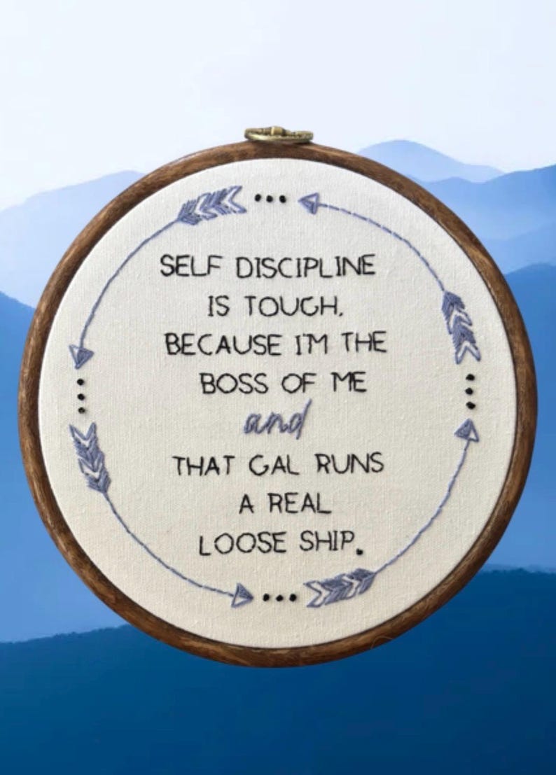 May include: Embroidered hoop art with the text "SELF DISCIPLINE IS TOUGH, BECAUSE I'M THE BOSS OF ME and THAT GAL RUNS A REAL LOOSE SHIP." The text is in black, with blue arrow and dot accents. The hoop is brown.