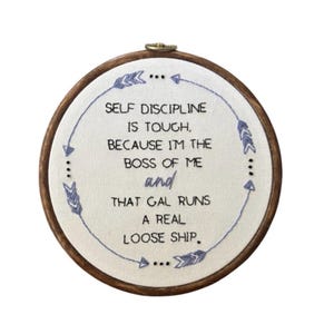 May include: An embroidered hoop featuring the text "SELF DISCIPLINE IS TOUGH, BECAUSE I'M THE BOSS OF ME and THAT GAL RUNS A REAL LOOSE SHIP." The text is in blue thread on white fabric, set within a wooden hoop.