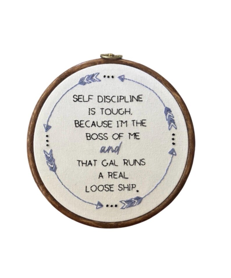 May include: An embroidered hoop featuring the text "SELF DISCIPLINE IS TOUGH, BECAUSE I'M THE BOSS OF ME and THAT GAL RUNS A REAL LOOSE SHIP." The text is in blue thread on white fabric, set within a wooden hoop.