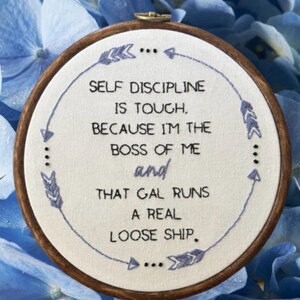 May include: A circular embroidery hoop with the embroidered quote: "SELF DISCIPLINE IS TOUGH, BECAUSE I'M THE BOSS OF ME and THAT GAL RUNS A REAL LOOSE SHIP." The design features blue stitching and is set against a blue floral backdrop.