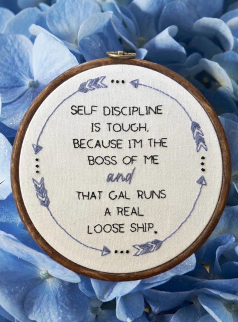 May include: A circular embroidery hoop with the embroidered quote: "SELF DISCIPLINE IS TOUGH, BECAUSE I'M THE BOSS OF ME and THAT GAL RUNS A REAL LOOSE SHIP." The design features blue stitching and is set against a blue floral backdrop.