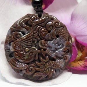 Pode incluir: Um pingente redondo de jade marrom e roxo com um design de dragão e fênix esculpido. O pingente está em um cordão preto e está descansando em uma flor rosa.