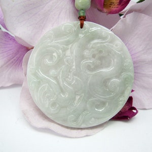 Pode incluir: Um pingente redondo de jade branco com um design de dragão esculpido. O pingente está pendurado em um cordão marrom.
