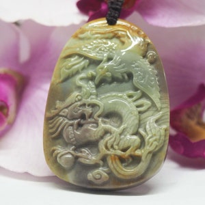 Pode incluir: Um pingente de jade esculpido com um design detalhado de um dragão e uma fênix. O pingente é de cor verde claro com detalhes marrons e brancos.