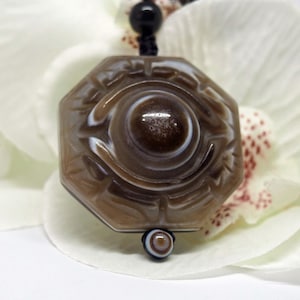 May include: Octagonal brown agate pendant with a white and brown eye design. The pendant is on a black cord and is surrounded by white flowers.
