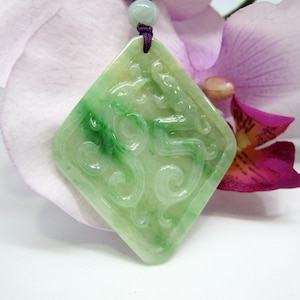 Pode incluir: Um pingente de jade verde com entalhes intrincados em forma de diamante. O pingente está em um cordão roxo e está rodeado de flores cor-de-rosa.