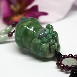 Puede incluir: Un colgante de jade verde tallado en forma de cerdo durmiendo. El colgante está unido a un cordón negro con cuentas rojas y blancas.