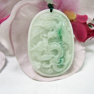 Pode incluir: Um pingente de jade branco esculpido com um design detalhado de um dragão e uma fênix. O pingente tem formato oval e tem um detalhe em jade verde.