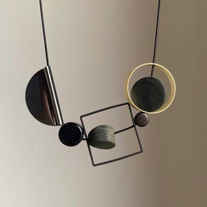 May include: A necklace featuring a black cord strung with various geometric shapes, including a dark metallic semicircle, a black circle, a thin black square frame containing a gray cylinder, a small dark bead, and a gold-toned circle with a black disc.