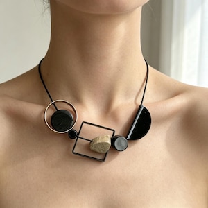 May include: A necklace featuring a black cord strung with geometric elements including a gold-toned circle, a black square frame, a black semi-circle, and several black circular beads, accented by a single textured, light-colored stone bead.