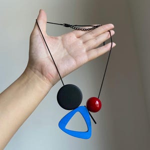 May include: A necklace featuring a black cord with a matte black circular pendant, a glossy red spherical bead, and a bright blue triangular frame pendant. The necklace includes a black metal chain extender with a lobster claw clasp closure.