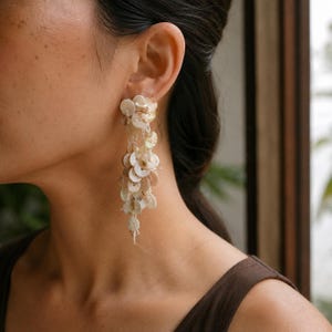 May include: A long, dangling earring composed of numerous small, circular, off-white shell discs attached to thin, light-colored threads. The discs are arranged in a cascading, layered cluster that hangs vertically from the earlobe.