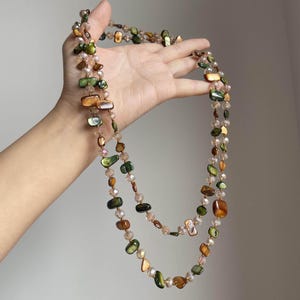 May include: A layered necklace featuring a mix of iridescent, green, and brown shell-shaped beads, interspersed with small, clear, faceted beads. The necklace is held up, showcasing its length and design. The beads have a natural, organic appearance.