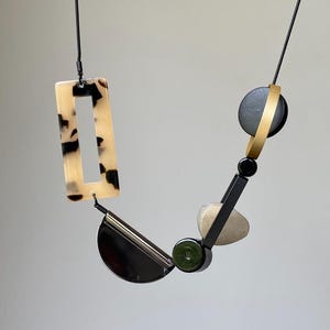 May include: A necklace featuring a black cord strung with various geometric shapes, including a rectangular tortoiseshell pendant, a black semi-circle, a dark green bead, a matte beige pebble-like piece, and a black circle encircled by a gold ring.