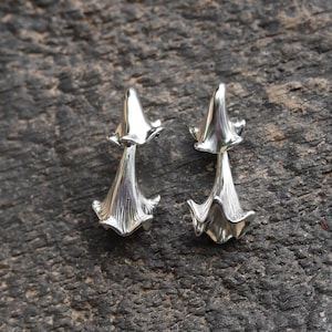 May include: A pair of silver-toned earrings shaped like bell-shaped flowers. Each earring features a smooth, rounded top section and a flared, fluted base with vertical ridges, reflecting light with a polished, metallic finish.