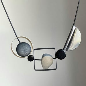 May include: A necklace featuring a black cord strung with geometric elements including a silver circle containing a dark gray sphere, a black square frame holding a light gray stone, two small black beads, and a polished silver crescent shape.